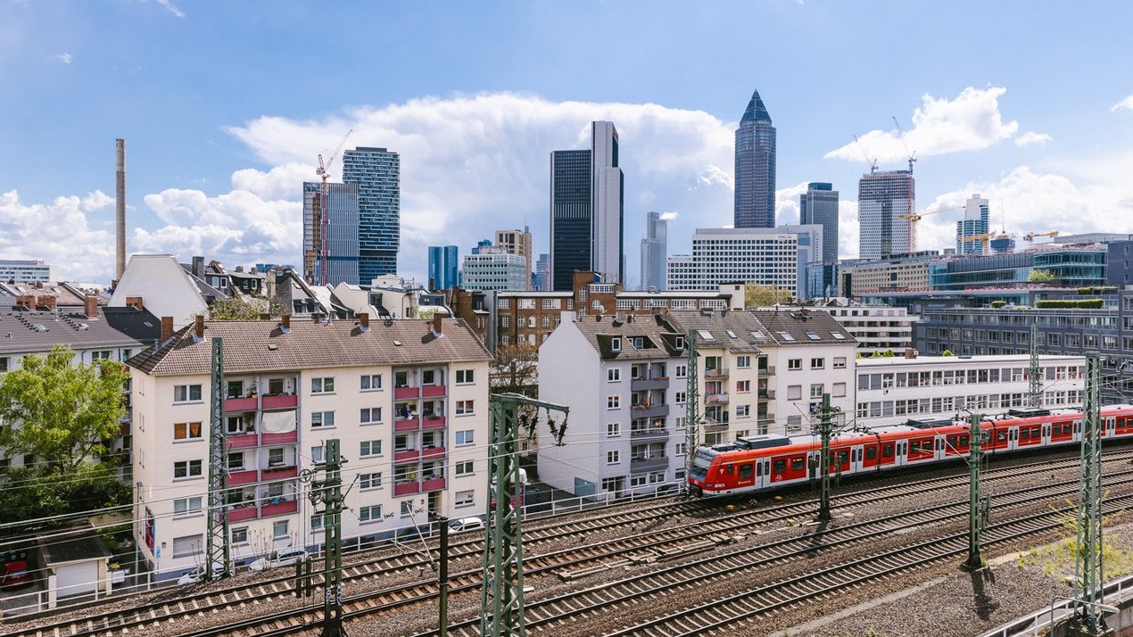 Ansicht von Frankfurt mit Wohnhäusern im Vordergrund, Bahntrassen mit roter S-Bahn und der Frankfurter Skyline mit Hochhäusern unter blauem Himmel.