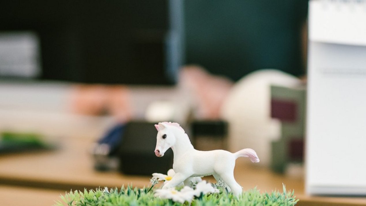 Kleine weiße Pferdefigur steht auf künstlichem Gras auf einem Schreibtisch, im Hintergrund sind unscharf Bürogegenstände zu erkennen.