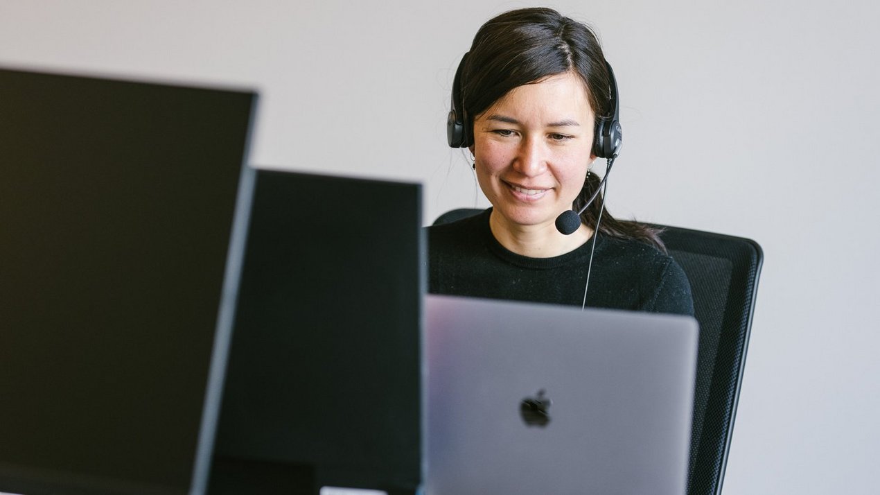 Frau mit Headset arbeitet am Laptop an einem Schreibtisch, blickt auf den Bildschirm und lächelt in einer modernen Büroumgebung.