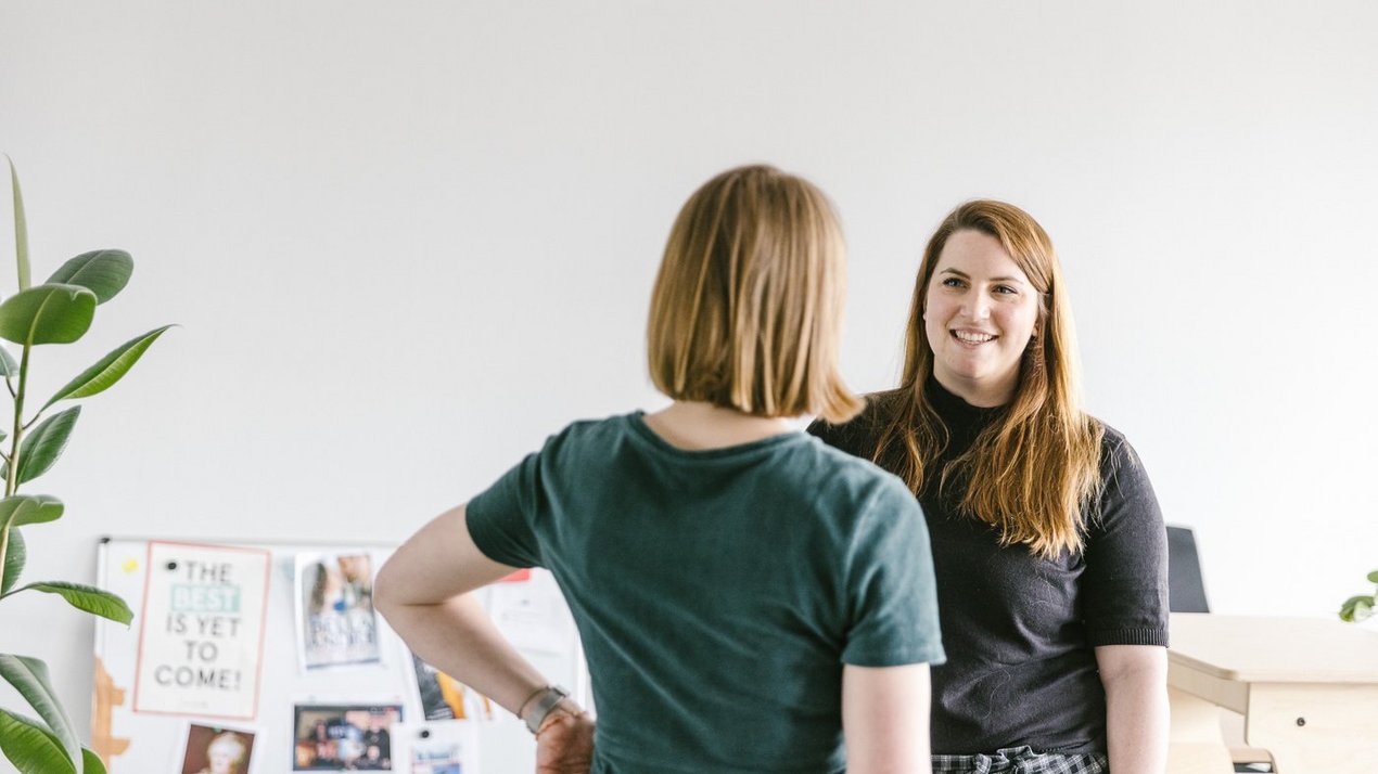 Zwei Frauen stehen sich in einem hellen Büro gegenüber und unterhalten sich entspannt, eine davon lächelt, im Hintergrund sind Pflanzen und eine Pinnwand zu sehen.