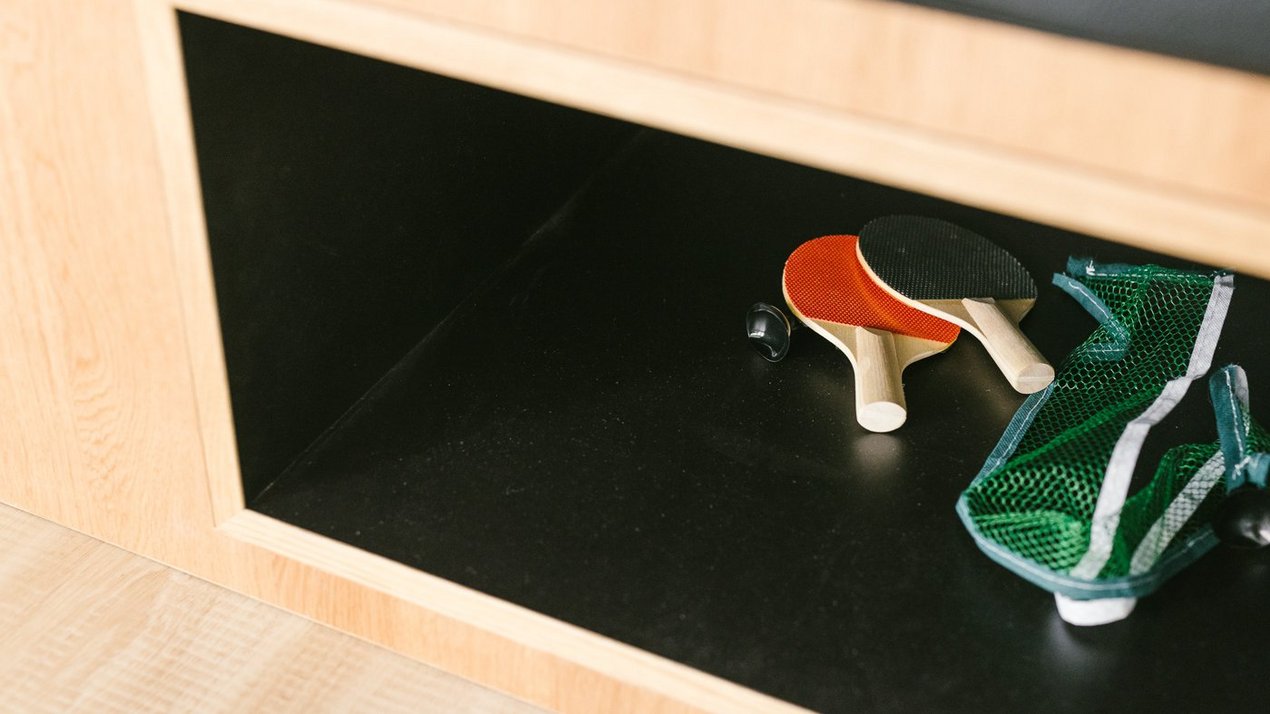 Offenes Regalfach mit Tischtennisschlägern, Ball und Netz, aufgeräumt in einem hellen Holzmöbel in einem Bürobereich..